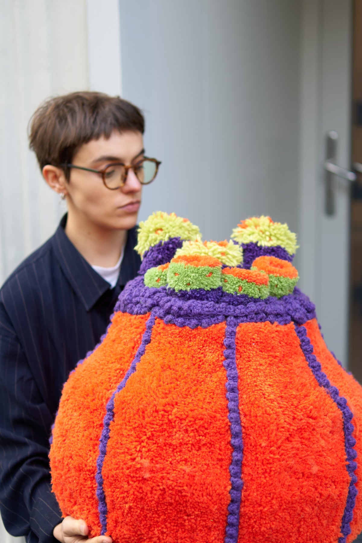 An upright, gourd-shaped textile sculpture featuring bright orange tufted vertical stripes. The top is crowned with textured, ruffled layers of purple and lime green wool. The artist is shown holding the piece in front of a modern storefront.